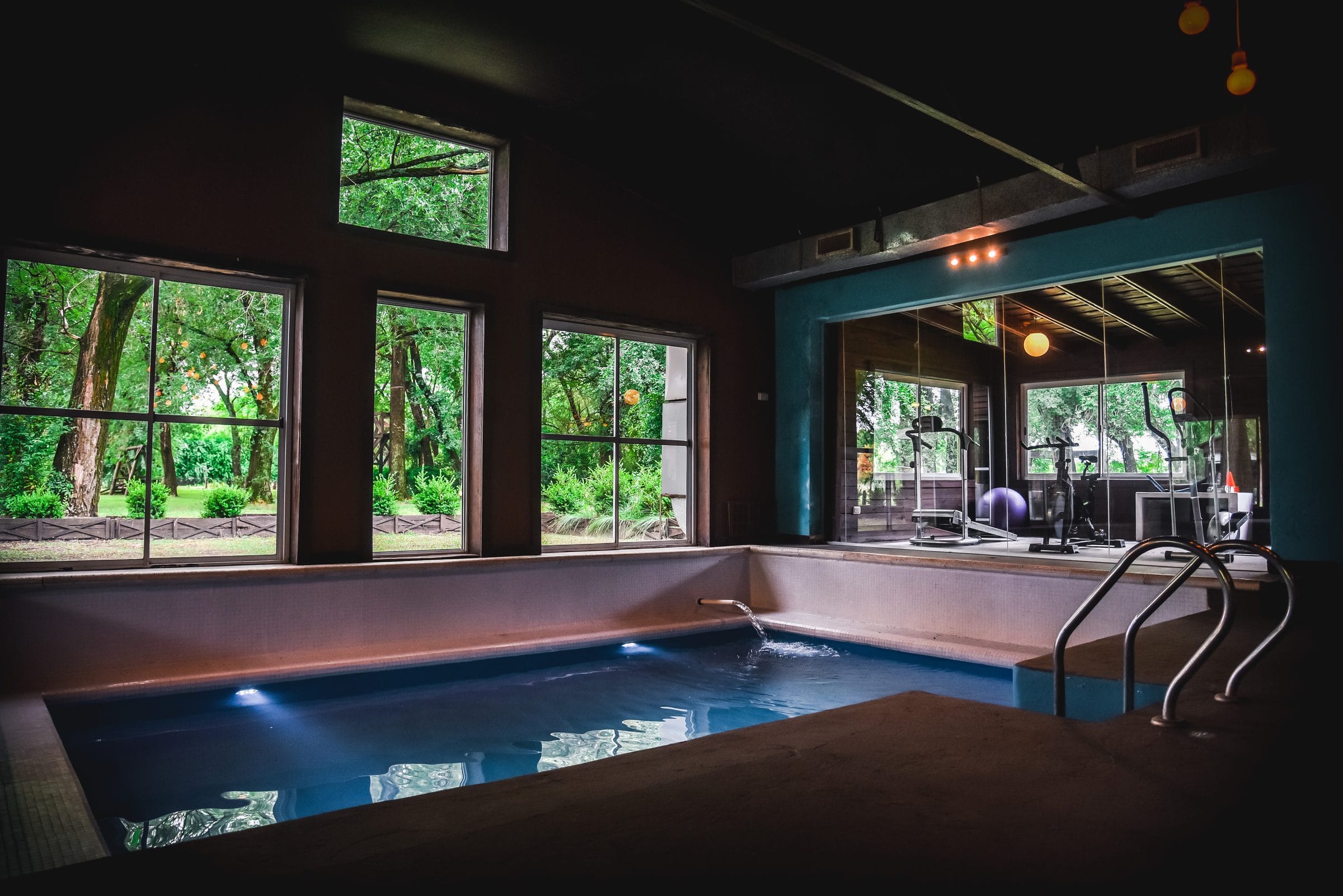 Pileta cubierta climatizada del Lagom Spa Rural: agua turquesa en calma bajo un techo de madera, con ventanales de piso a techo que dan al parque.
